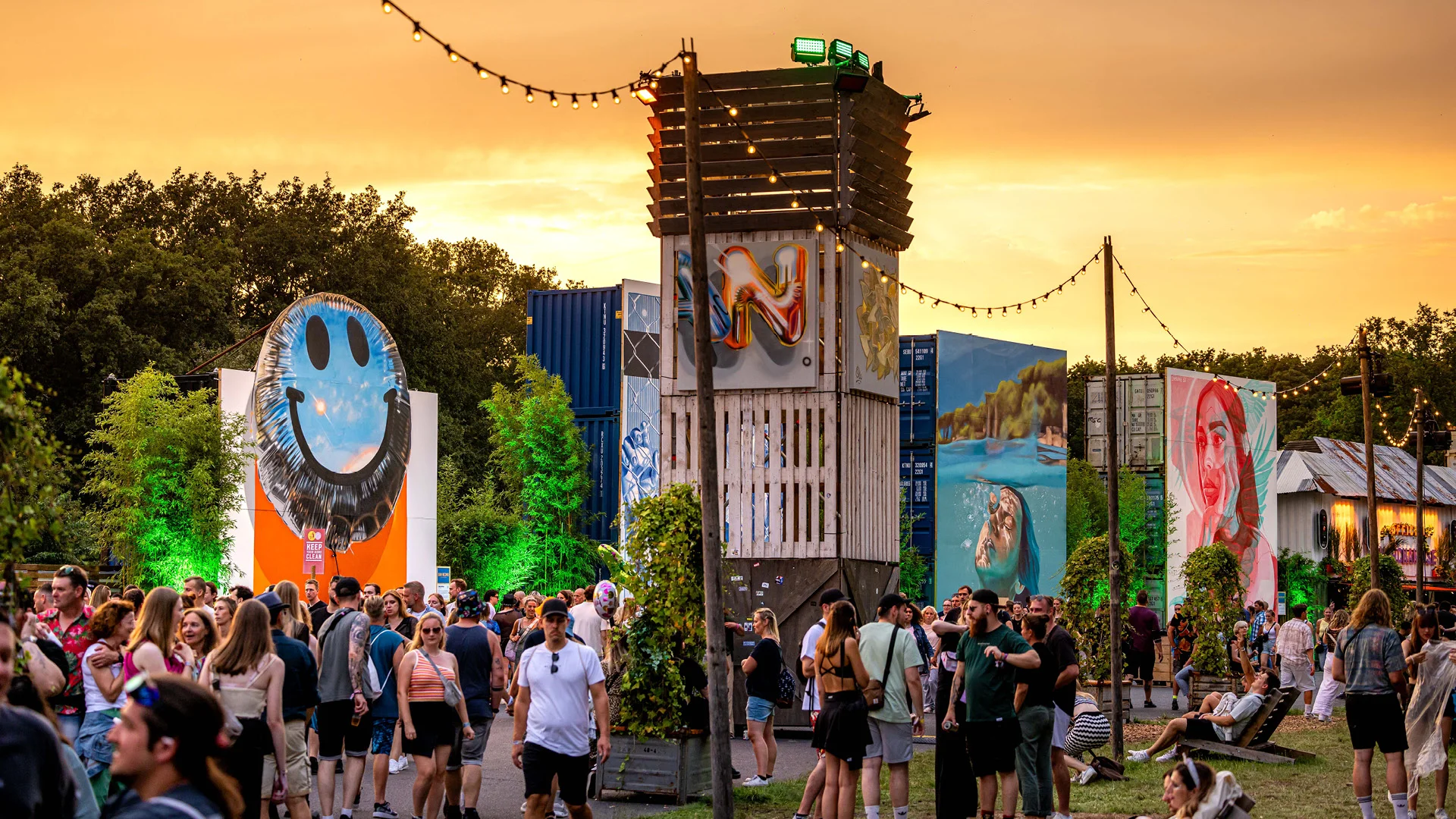 Weitwinkelaufnahme des Urban Art Districts bei Sonnenuntergang mit mehreren bemalten Containern, darunter ein riesiger Smiley und Porträts in bunten Farben.