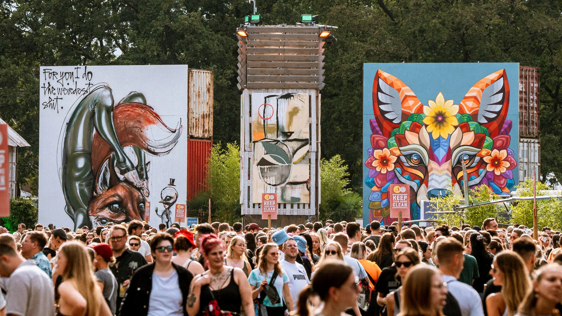 Drei großformatige Kunstwerke im Urban Art District des San Hejmo Festivals: links ein surrealistischer Fuchs mit Handstand-Figur, mittig ein abstraktes Bild, rechts ein farbenfrohes, ornamentales Fuchsporträt. Davor zahlreiche Festivalbesucher.