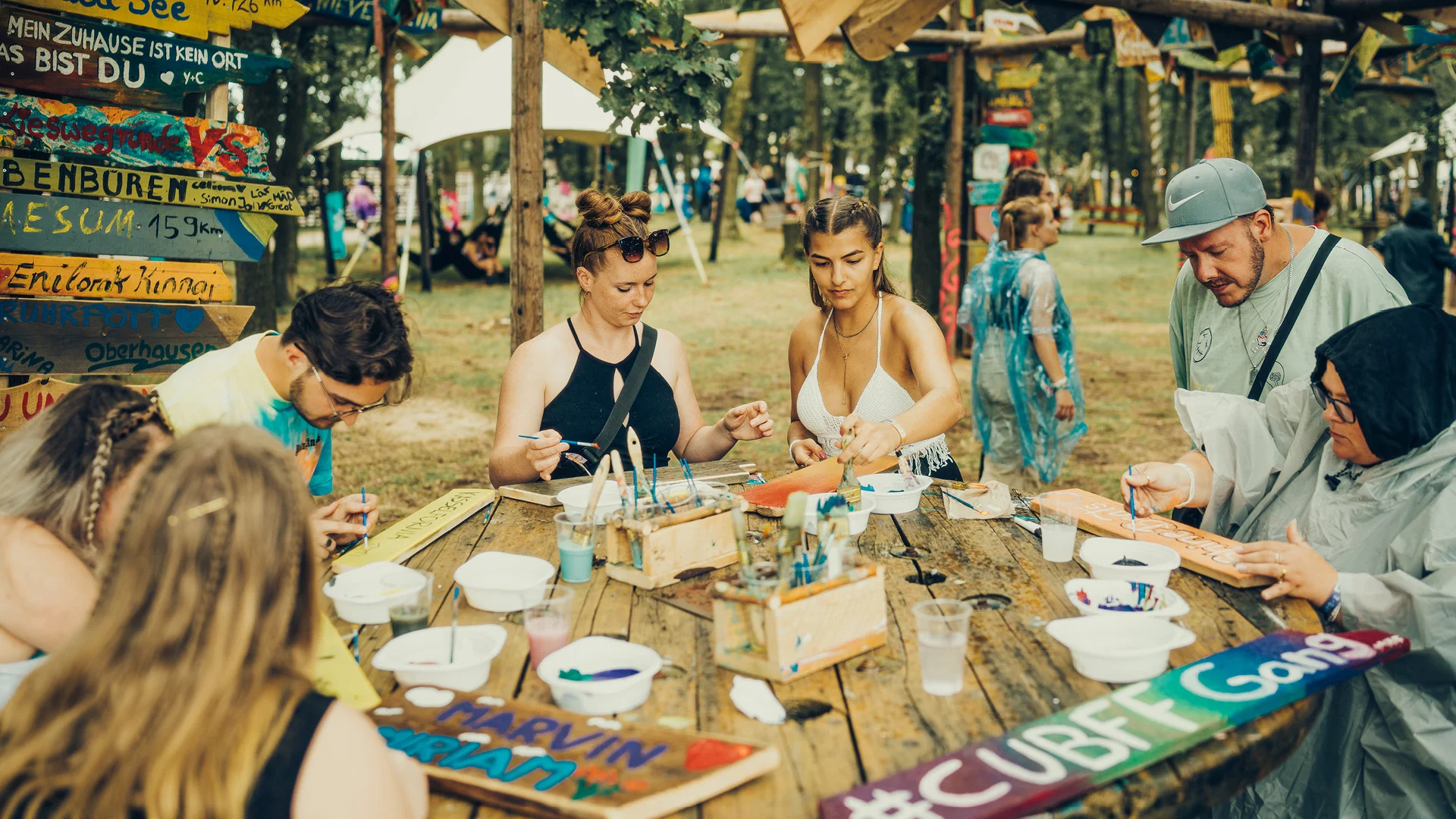 Teilnehmer:innen des „Schilderwald“-Workshops beim San Hejmo Festival bemalen gemeinsam bunte Holzschilder an einem großen Tisch im Freien.