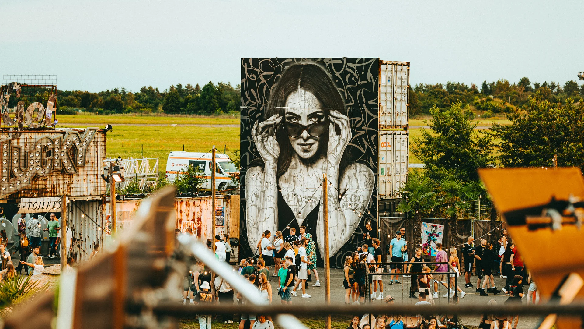 Hyperrealistisches Wandgemälde einer Frau mit Sonnenbrille auf einem hohen Container, umgeben von kalligrafischem Hintergrund. Davor Festivalbesucher im Urban Art District des San Hejmo Festivals.