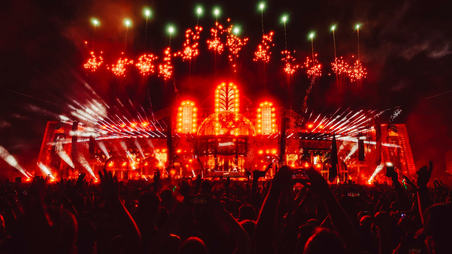 PAROOKAVILLE Mainstage 2024 bei Nacht in rotes Licht getaucht, mit intensiver Pyroshow, Feuerwerk und Scheinwerfern – die ikonische Kuppel strahlt hell, während die Crowd im Vordergrund jubelt, tanzt und den Moment mit Handys festhält.