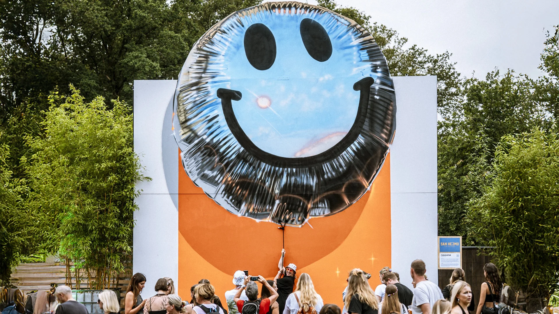 Täuschend echt wirkende Kunstinstallation in Form eines riesigen Luftballons mit Smiley-Gesicht vor orange-weißem Hintergrund, umringt von Festivalbesuchern im San Hejmo Urban Art District.